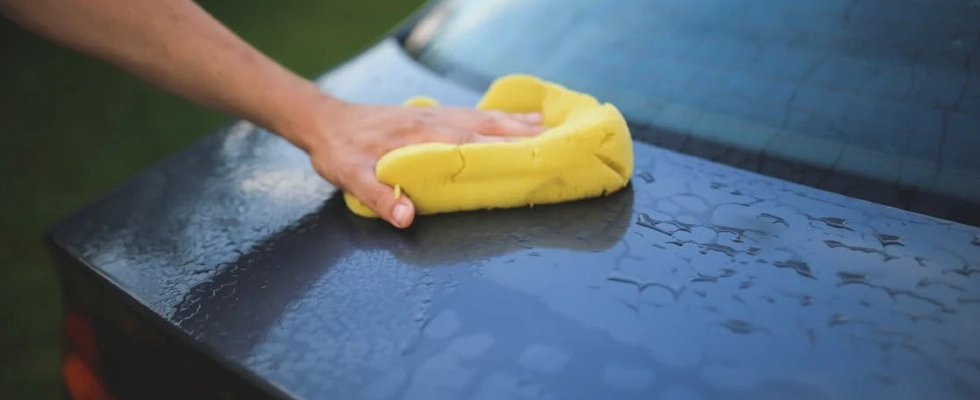 Hand cleaning car with a yellow sponge, showcasing car detailing and washing process.