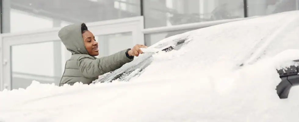 woman removing snow from the front windshield of her car.